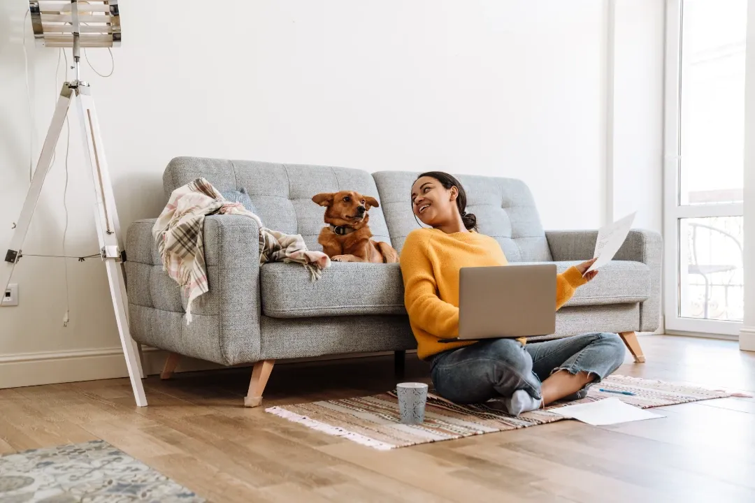 Woman sitting on the floor with a laptop, smiling at a brown dog on a gray sofa in a bright living room