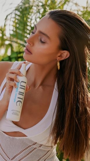 Woman spraying UNITE hair product on long brown hair, showcasing smooth, shiny styling results.
