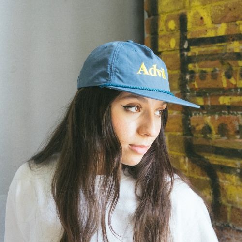 Woman wearing light blue Advil snapback hat with embroidered logo indoors near brick wall.