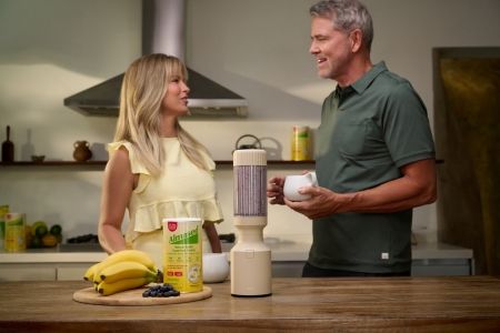 Couple enjoying protein smoothies in a modern kitchen with Almased nutrition shake mix and fresh fruit on the counter.