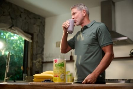 Man drinking a blueberry smoothie beside Almased protein powder, bananas, and blueberries in a bright kitchen.