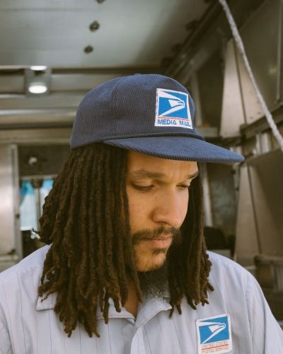 Man wearing navy USPS Media Mail inspired snapback hat in industrial setting.