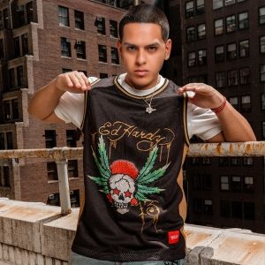 Man modeling a black Ed Hardy basketball jersey with skull and wing graphic in an urban rooftop setting.