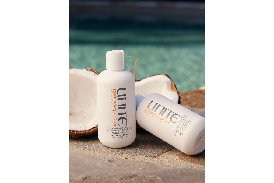 UNITE Boing curl cream and leave-in conditioner bottles on sand near pool with coconuts for tropical theme