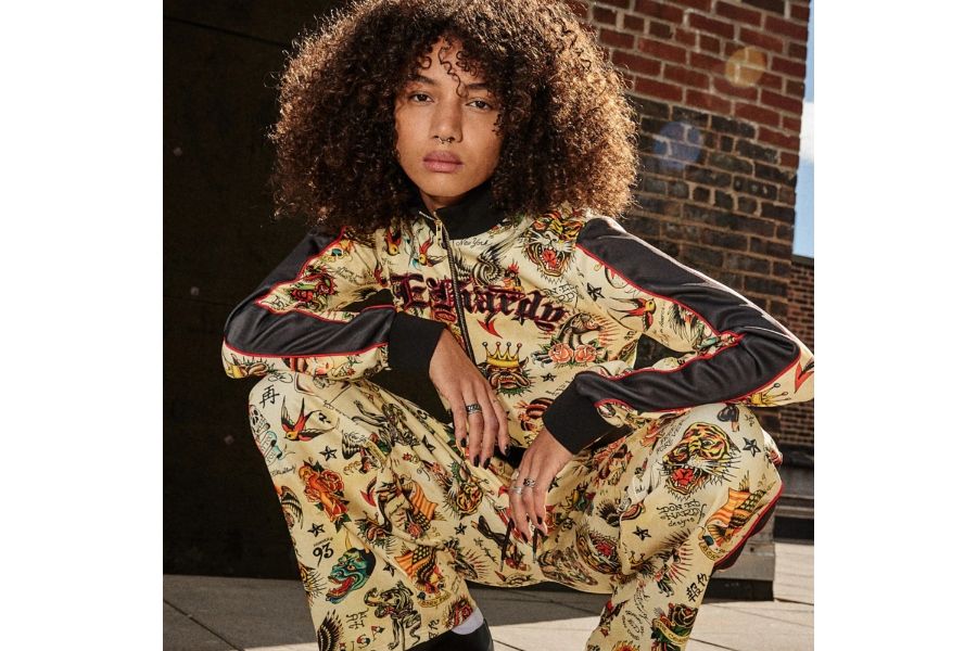 Person crouching in colorful Ed Hardy tracksuit with tattoo-style graphics of tigers, roses, crowns, and swallows