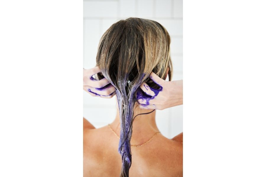 Back view of person applying UNITE Hair purple shampoo to wet blonde hair in bathroom with white tiled background