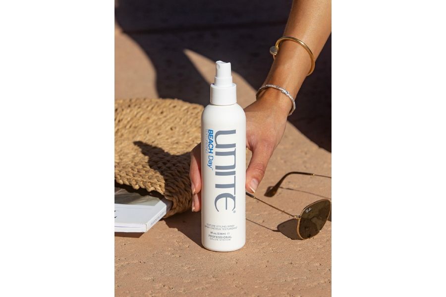 Hand with bracelets holding UNITE Hair Beach Day Texturizing spray outdoors with straw item, book, and sunglasses