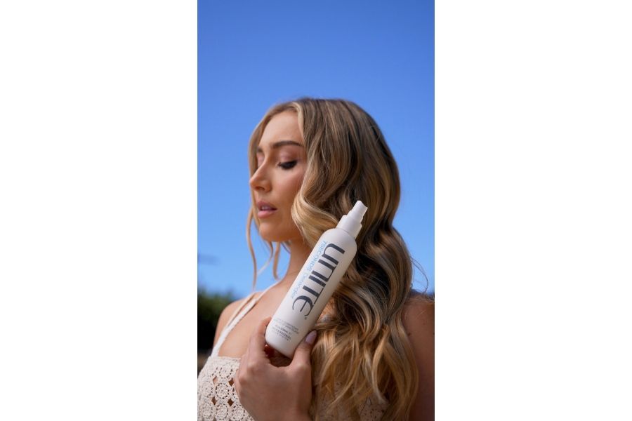 Person outdoors holding UNITE HAIR 7SECONDS Detangler leave-in conditioner bottle with long blonde hair.