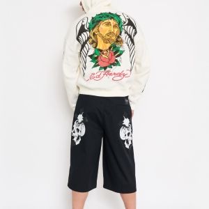 Back view of model wearing ivory Ed Hardy zip hoodie with large religious graphic and black long shorts with skull prints.