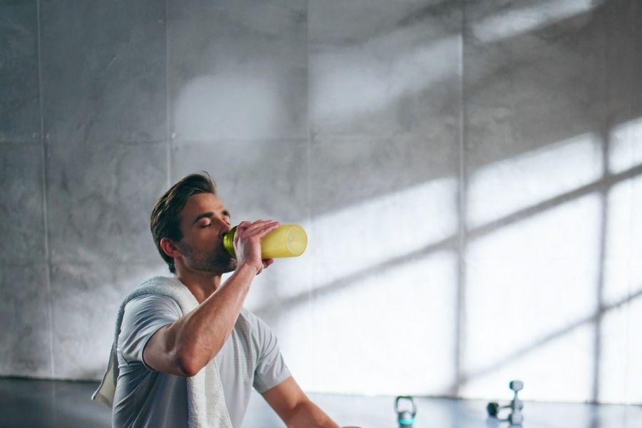 Almased person hydrating with yellow bottle on gym bench with towel and equipment, emphasizing fitness recovery and healthy lifestyle