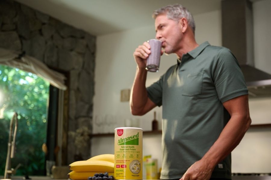 Almased person enjoying smoothie in kitchen with protein container, bananas and blueberries, showcasing balanced nutrition and wellness