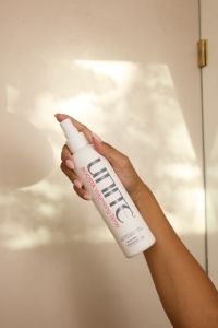 Hand holding a UNITE Boosta Volumizing Spray bottle against a softly lit beige background.