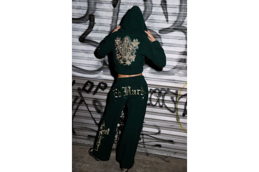 Person from behind in green sherpa Ed Hardy hoodie and pants with gold embroidery, against graffiti wall