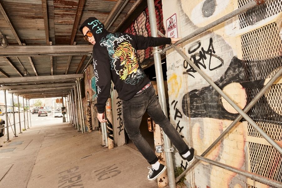 Person in black Ed Hardy hoodie with tiger back graphic leaning off scaffolding on a graffiti-covered street